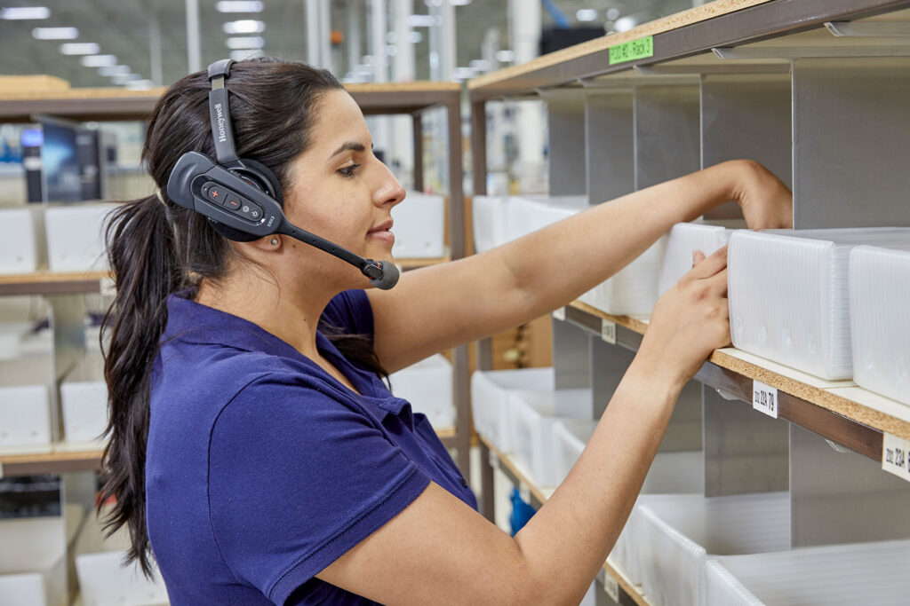 Pracownik odkładający towar na półkę używający urządzenia do komunikacji głosowej firmy Honeywell