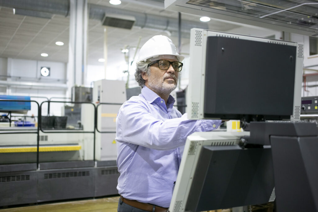 Serious male plant manager in hardhat and glasses operating industrial machine, pushing buttons on control panel. Copy space. Industrial technology or machinery concept