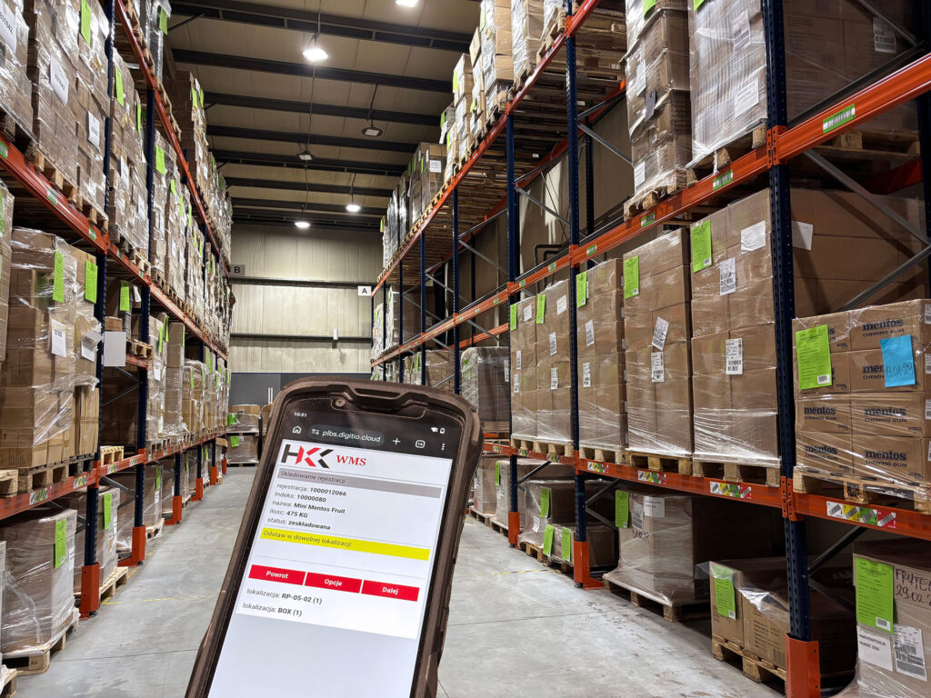 Komputer mobilny z systemem WMS, w tle regały magazynowe z paletami