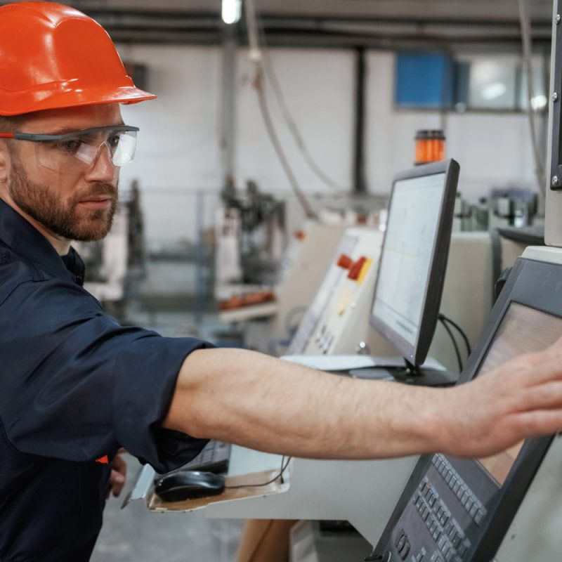 View from the side, working with machine, operator. Factory worker is indoors with hard hat.